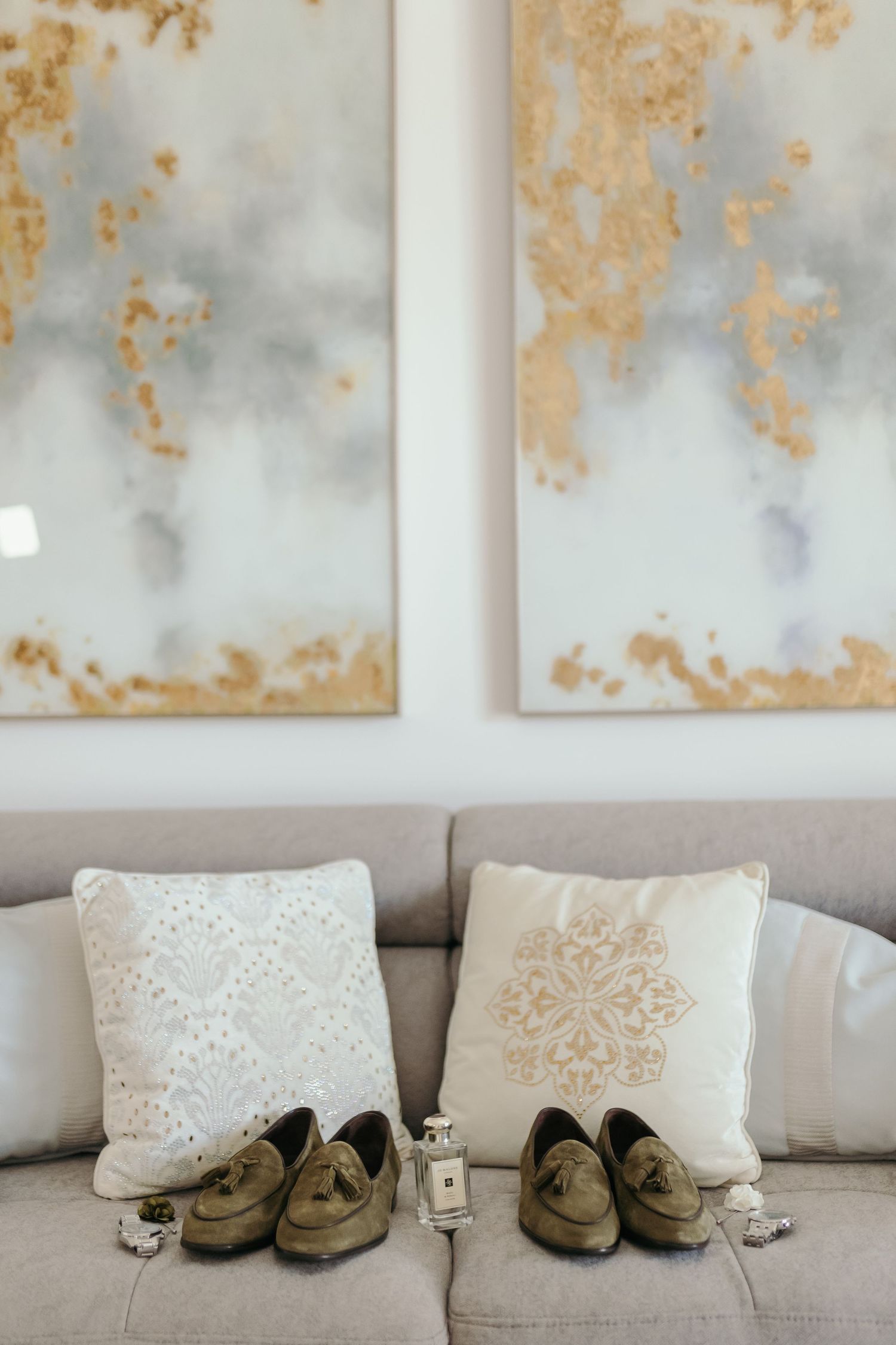 Two pairs of matching tan suede loafers on decorative pillows against gold artwork.