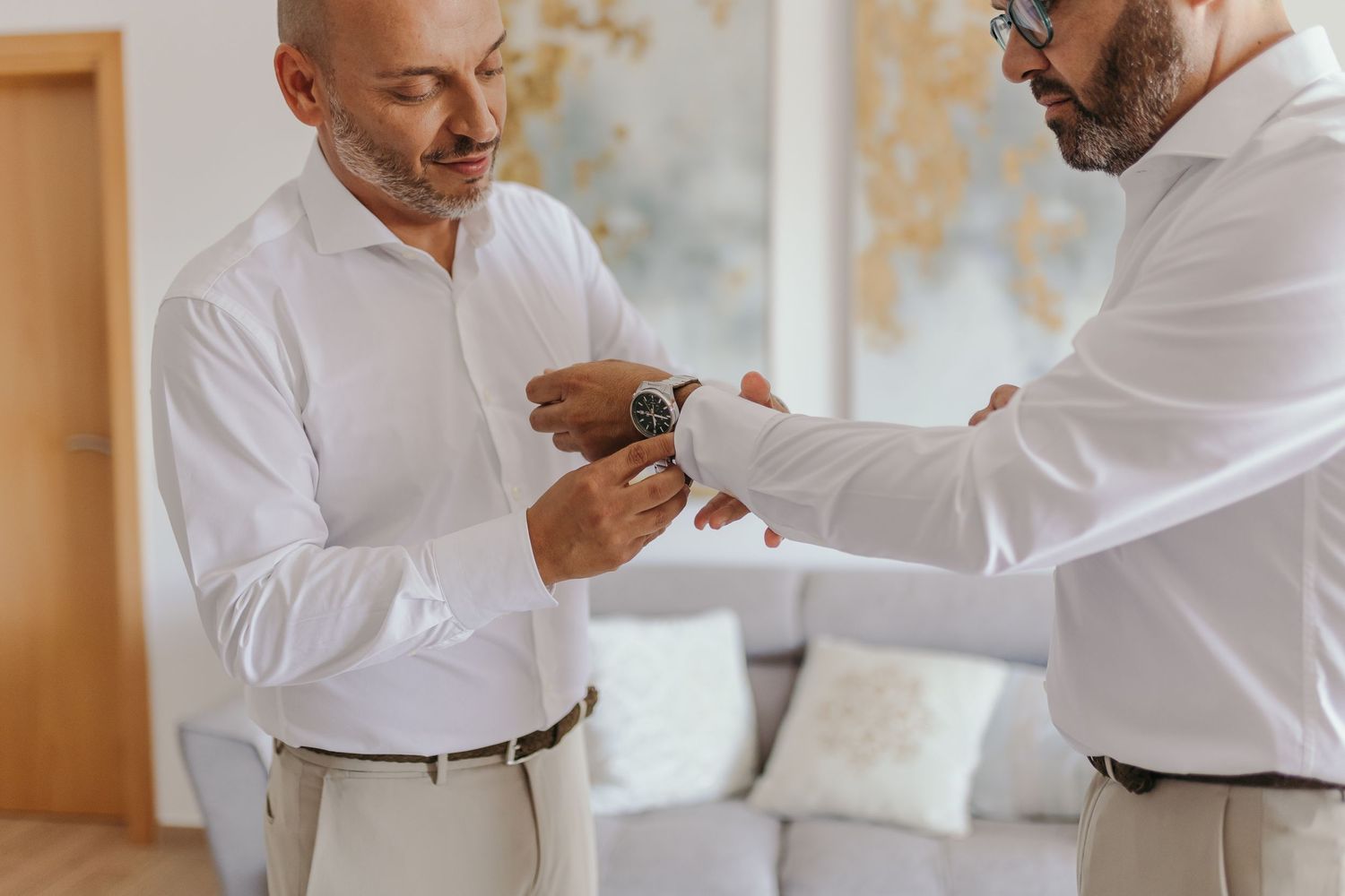 Two men in white shirts with one helping the other fasten a watch cufflink in a bright interior.