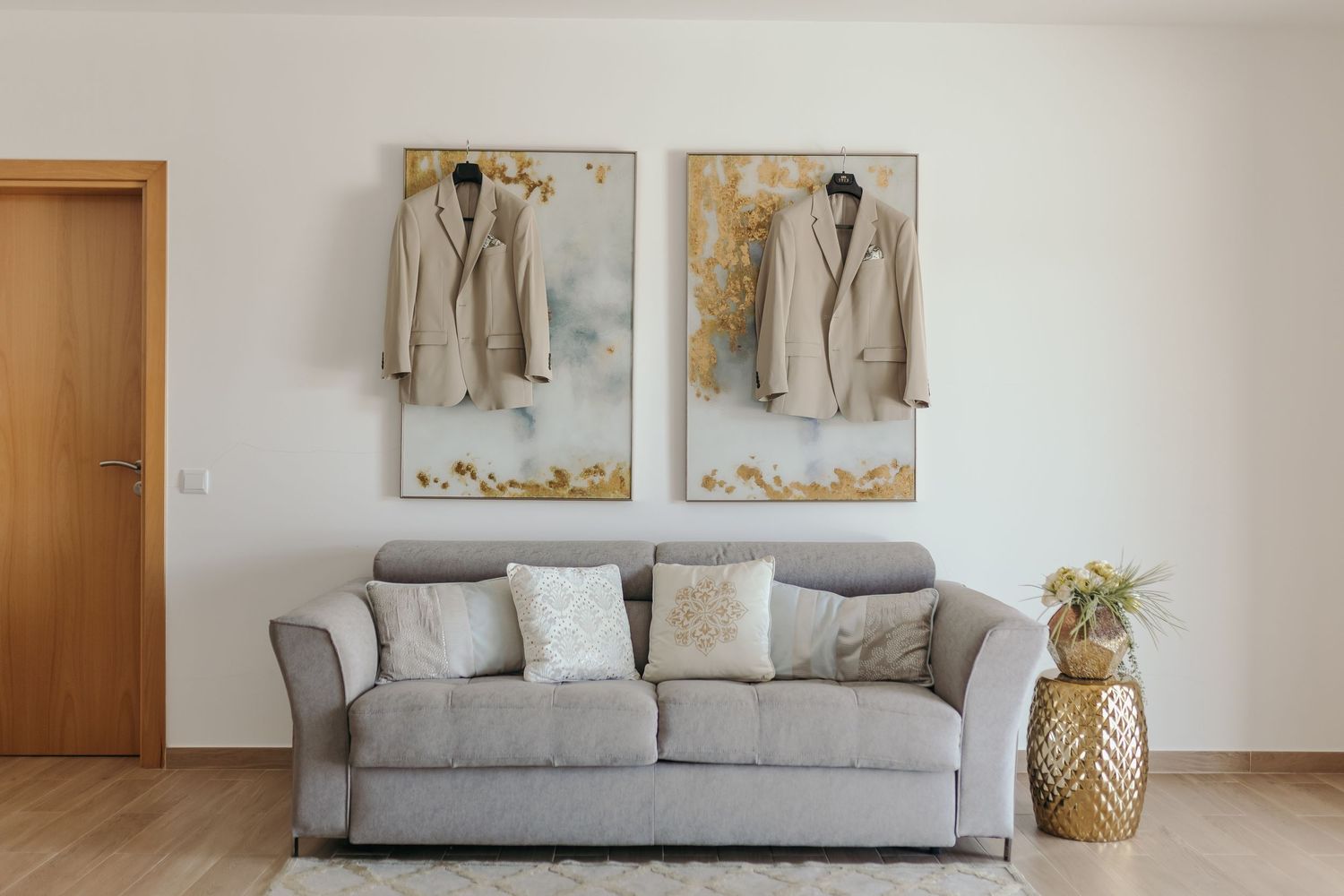Two matching beige wedding suits hanging over gold abstract artwork above gray sofa.