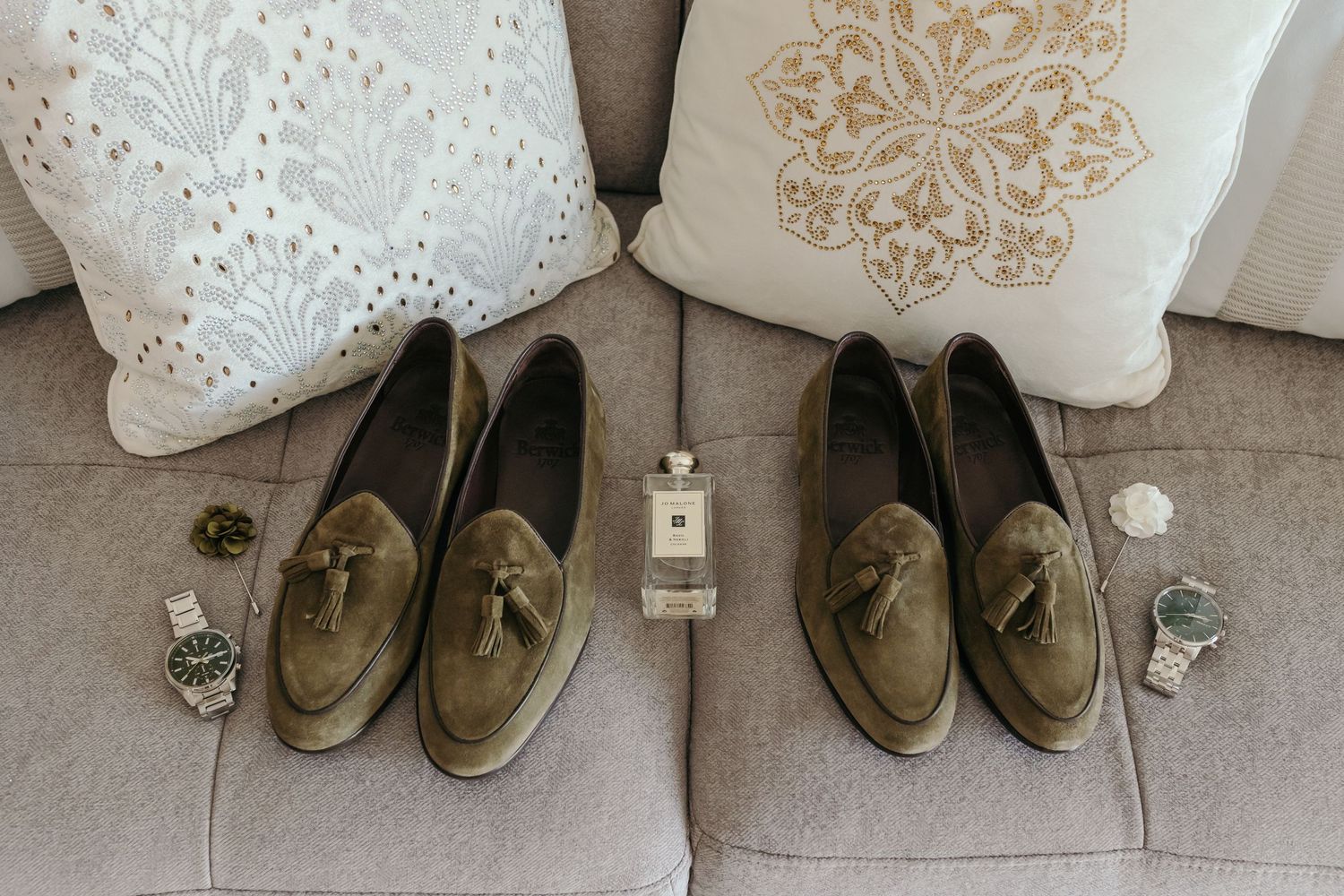 Matching pairs of tan suede loafers with wedding accessories on gray sofa.