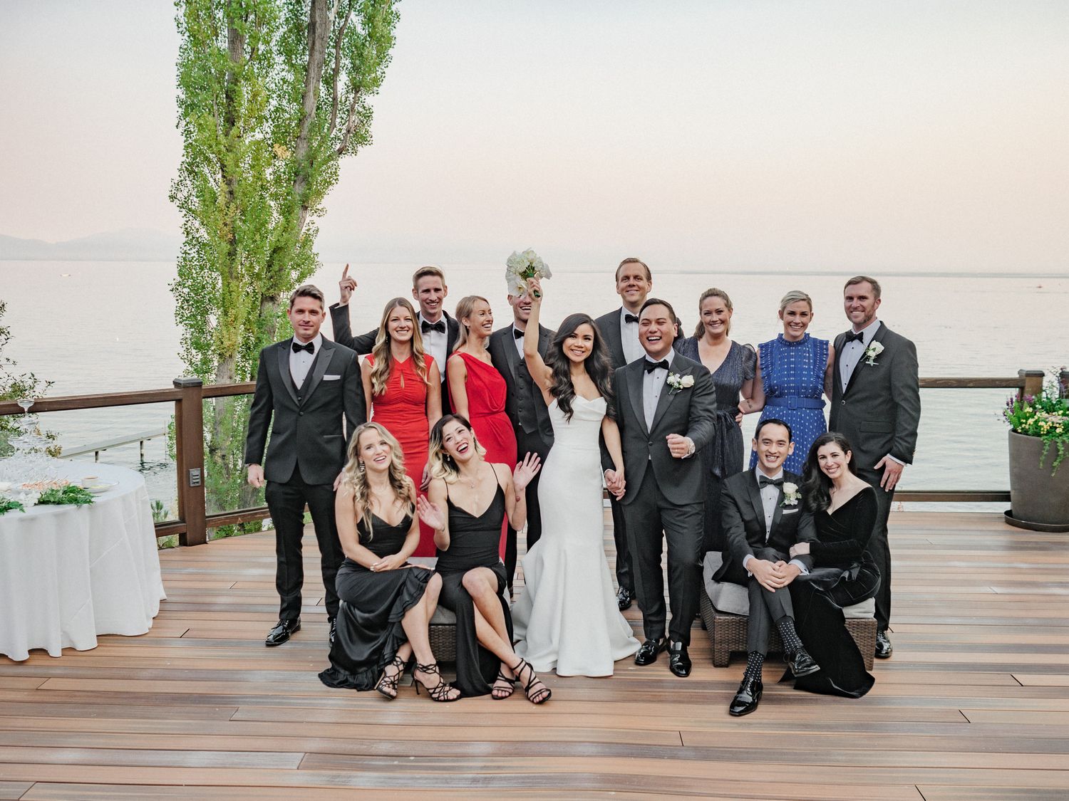 A wedding party poses together on a wooden deck overlooking a waterfront at sunset with tall trees in the background.