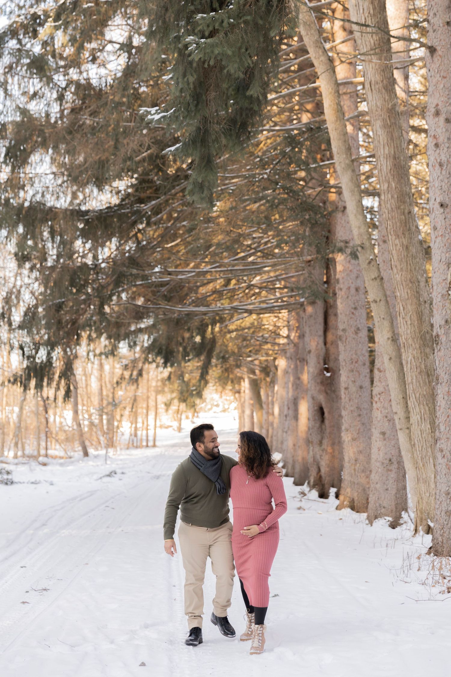 pregnant couple walking through the evergreen trees holding hands by Sheldon estate