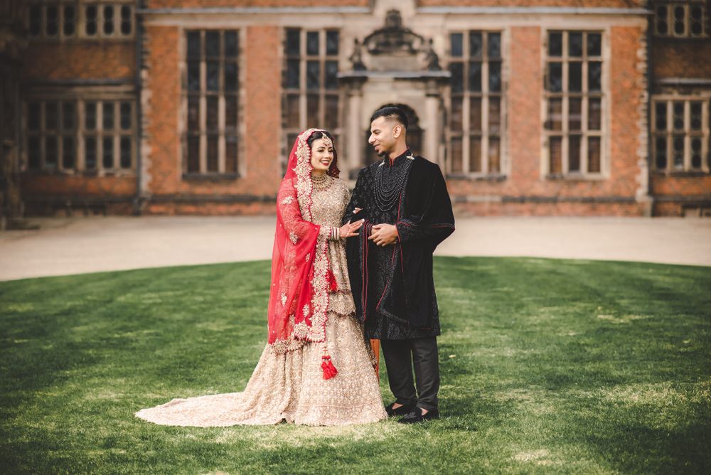 A couple in traditional South Asian wedding attire poses in front of a historic brick building.