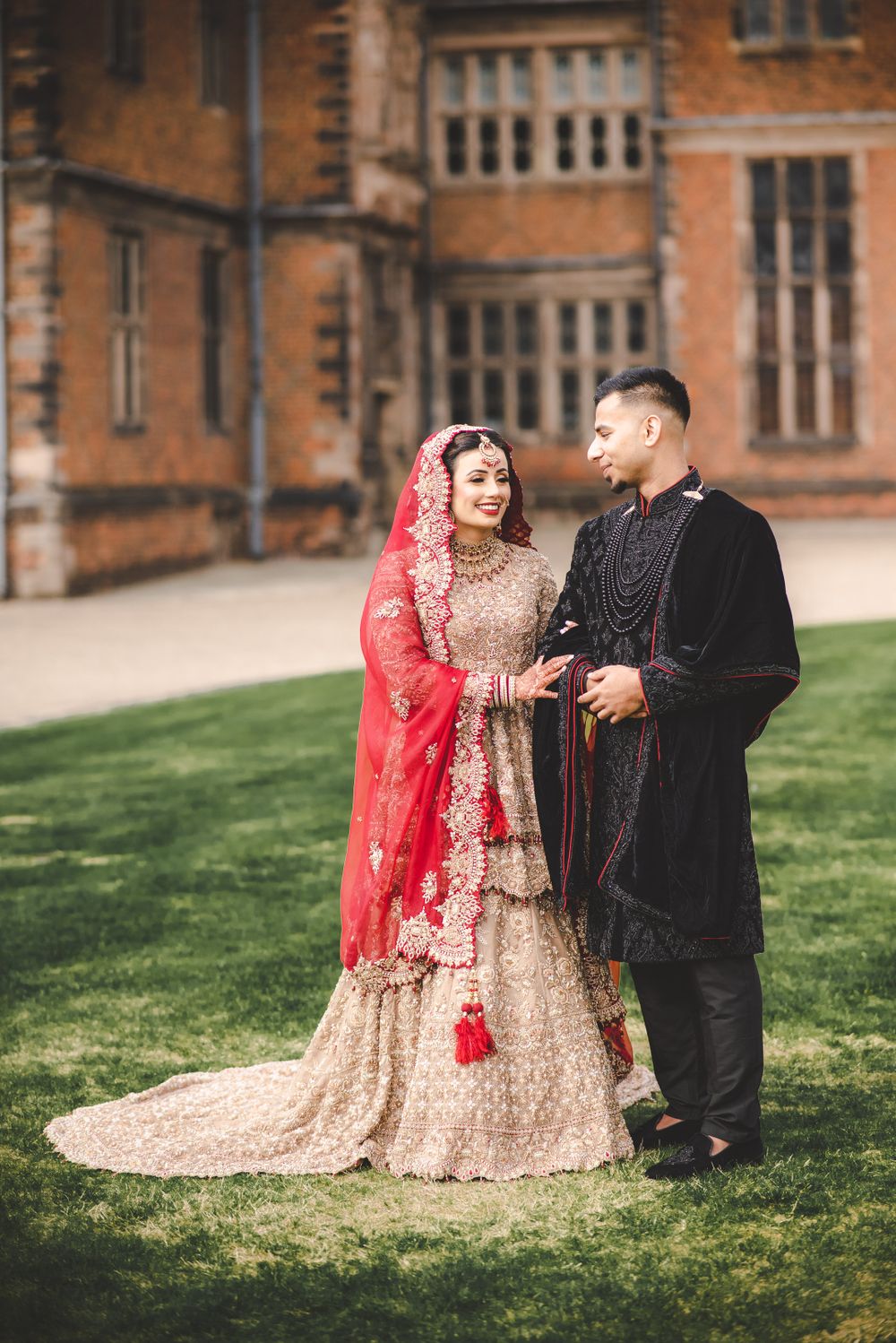 A couple in traditional South Asian wedding attire stands together on a manicured lawn.