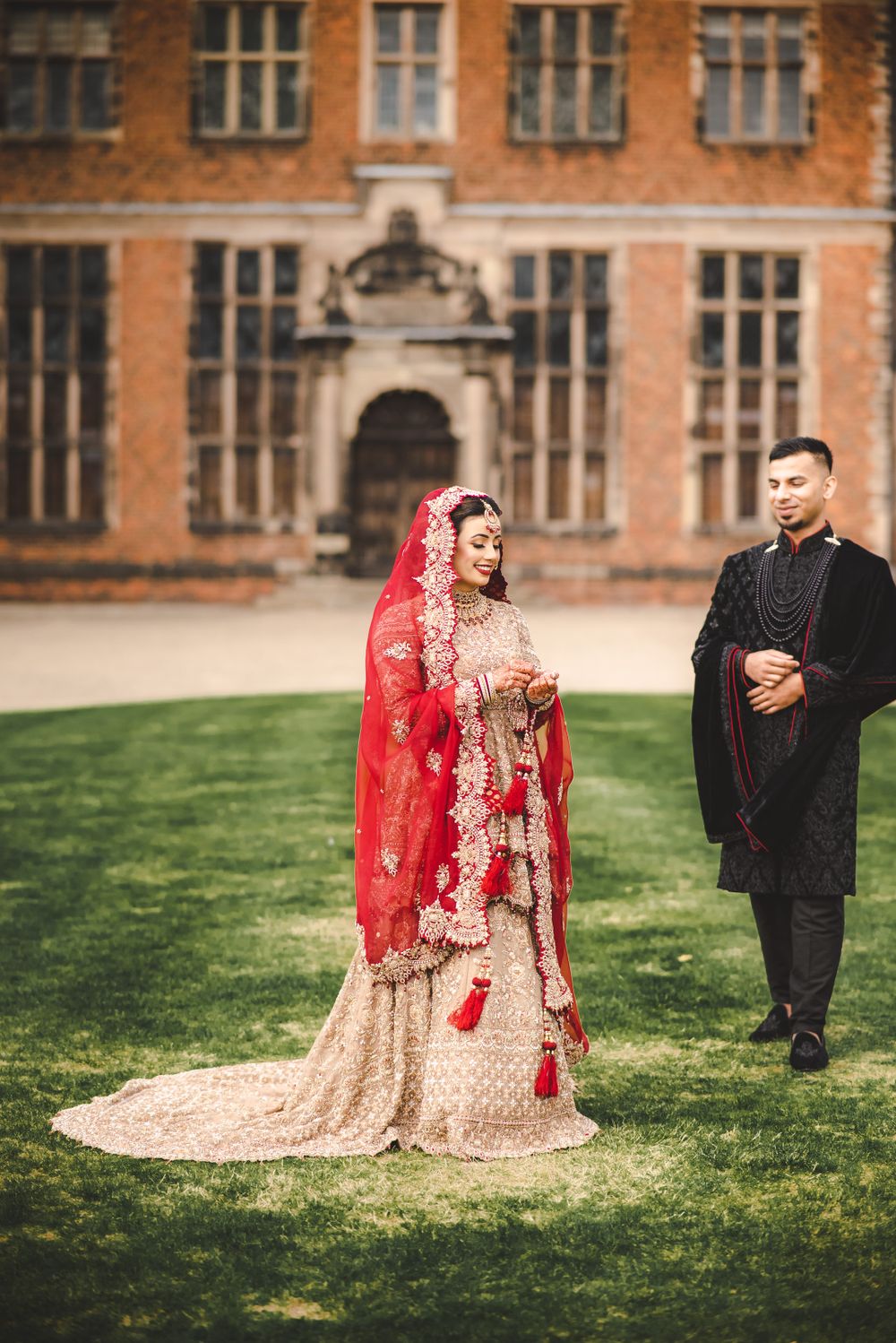 A bride in red and gold lengha stands with groom in front of historic architecture.