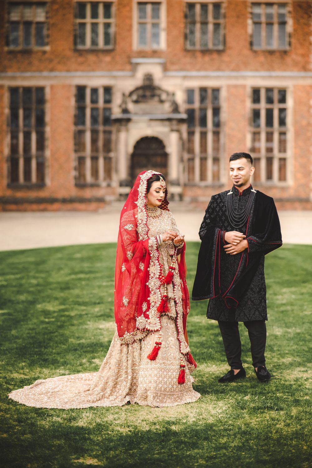 A bride in red and gold with groom in black pose on lawn of historic building.