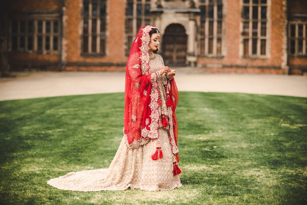 A stunning bridal portrait in traditional South Asian wedding attire with red dupatta against a historic building backdrop.