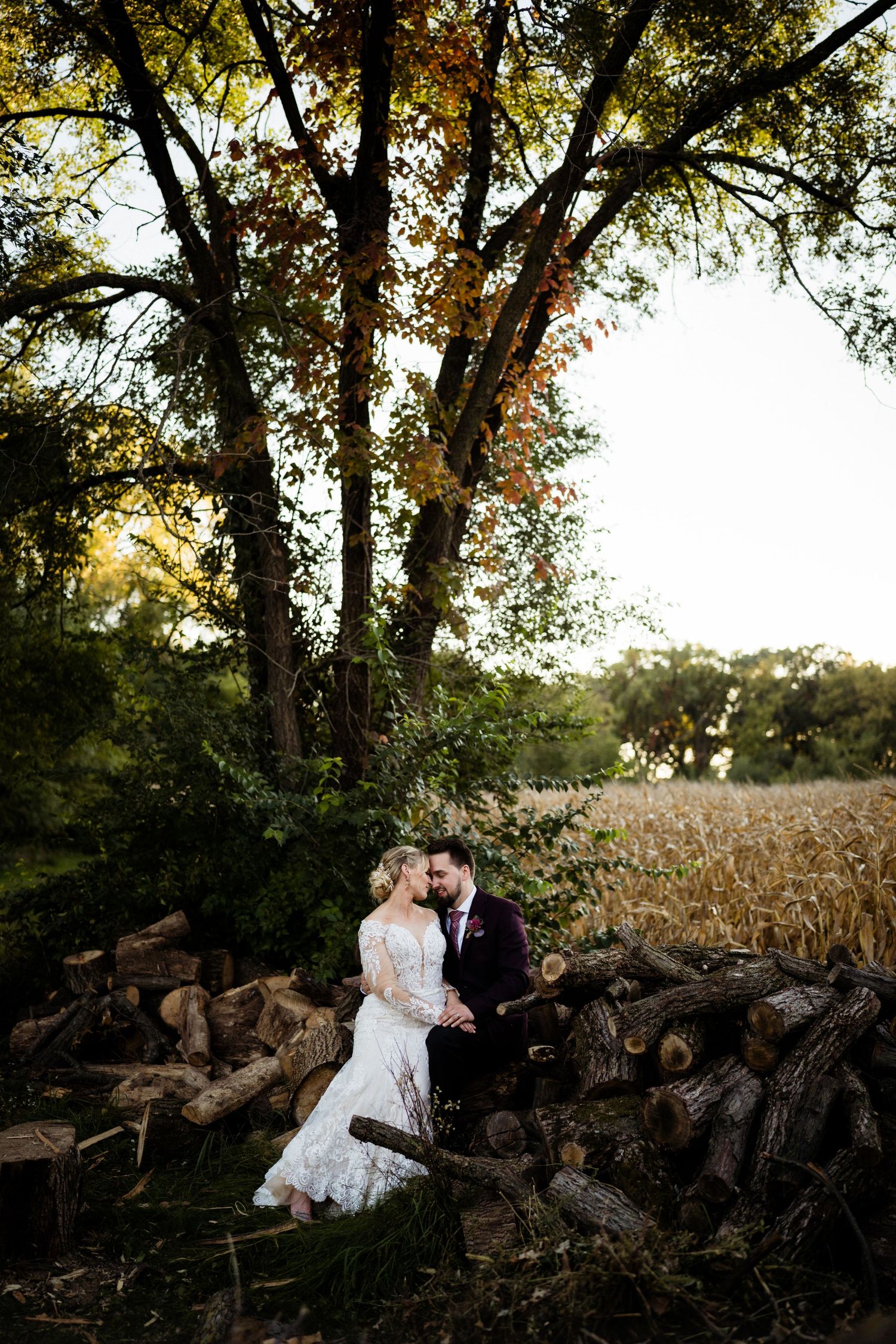 Romantic sunset portrait of a couple by a rustic woodpile surrounded by autumn trees.