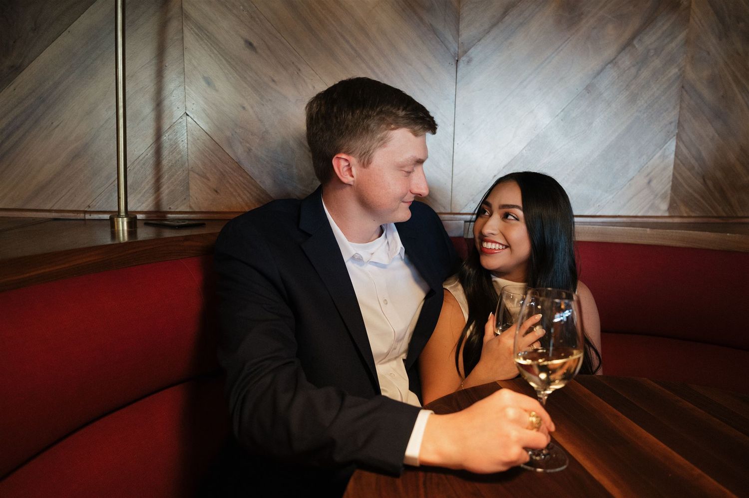 Couple shares an intimate moment while enjoying wine in a cozy restaurant booth with wooden paneled walls.