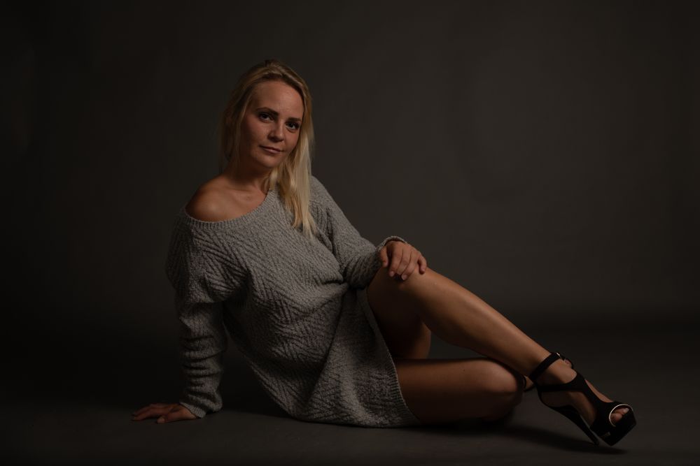 Blonde woman in grey oversized sweater posing on floor with crossed legs in dark studio.