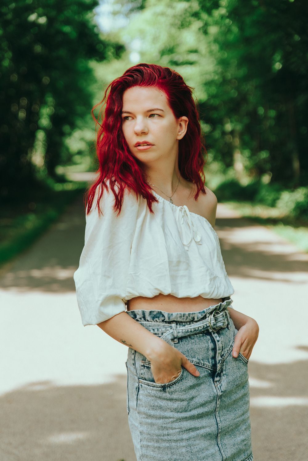 Young woman with vibrant red hair wearing white off-shoulder crop top and denim skirt standing on a tree-lined path.