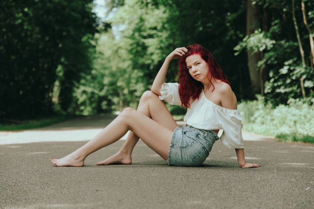 Red-haired woman in white off-shoulder top and denim skirt sitting barefoot on a sunlit forest road.