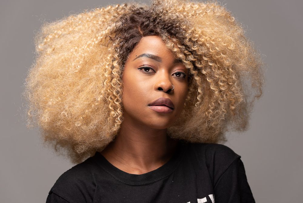 Young Black woman with large blonde afro hairstyle wearing black t-shirt against grey background.