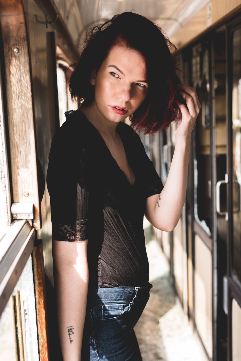 Dark-haired woman with tattoos in black lace top posing in vintage train corridor with moody lighting.