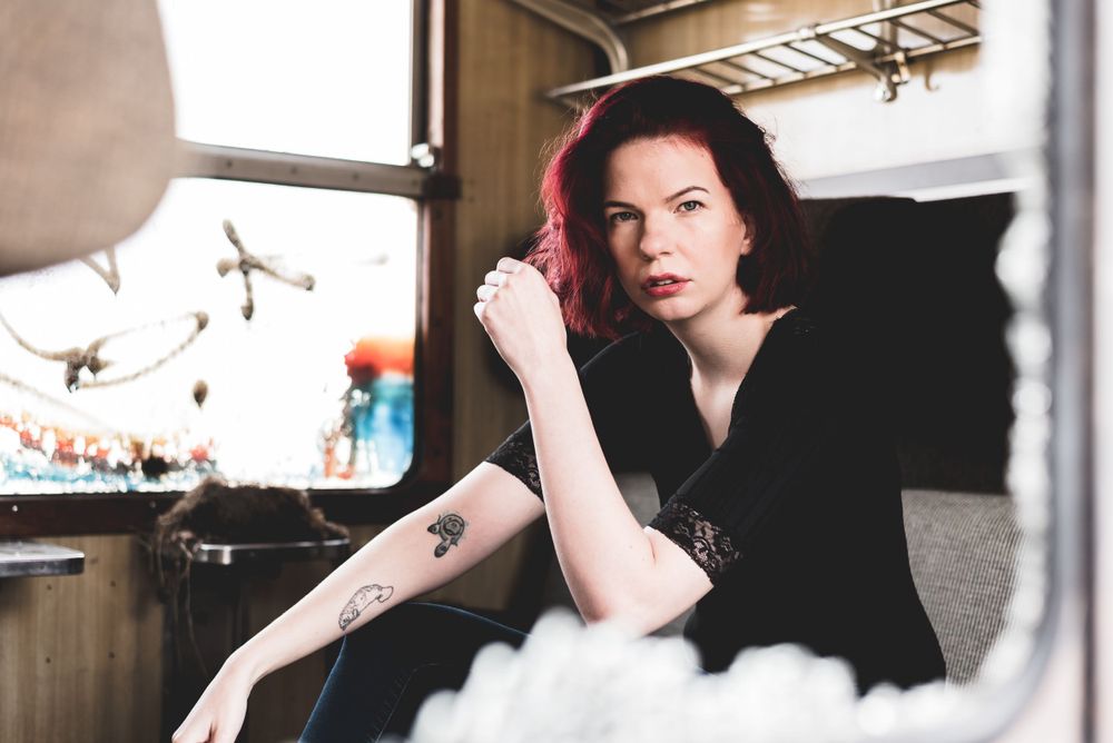 Tattooed red-haired woman sitting pensively in vintage train compartment with soft natural window light.