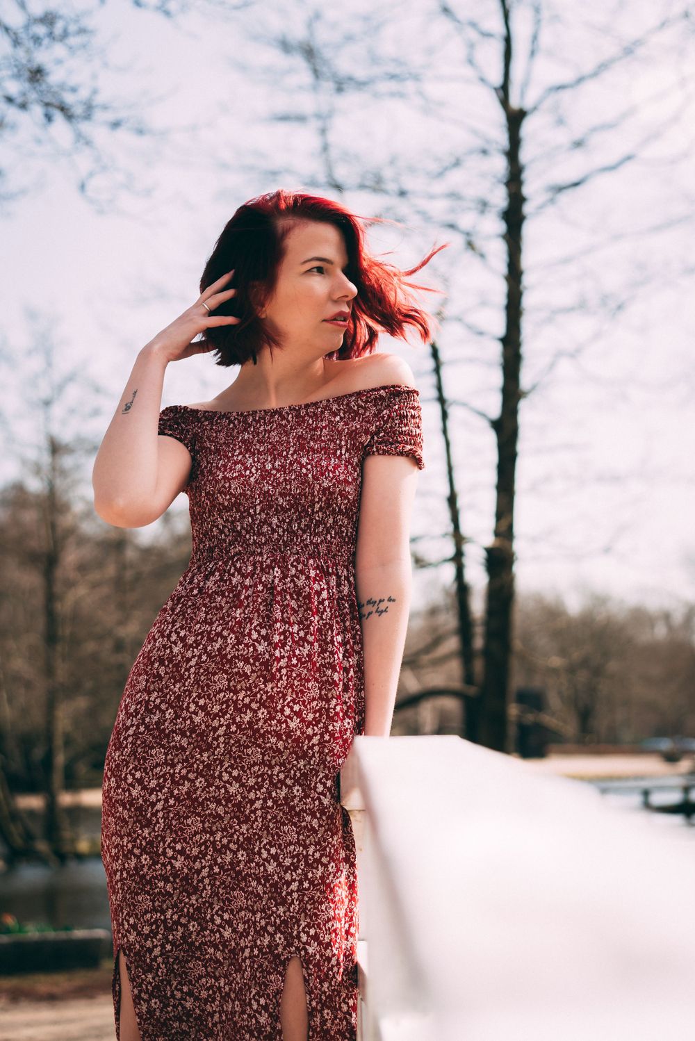 Short red-haired woman in floral off-shoulder maxi dress standing by white railing in outdoor park setting.
