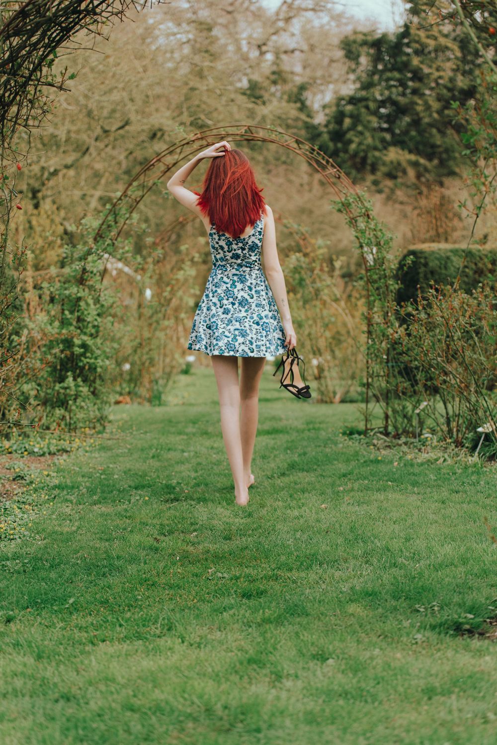 Red-haired woman in floral mini dress walking barefoot through garden archway carrying heels.