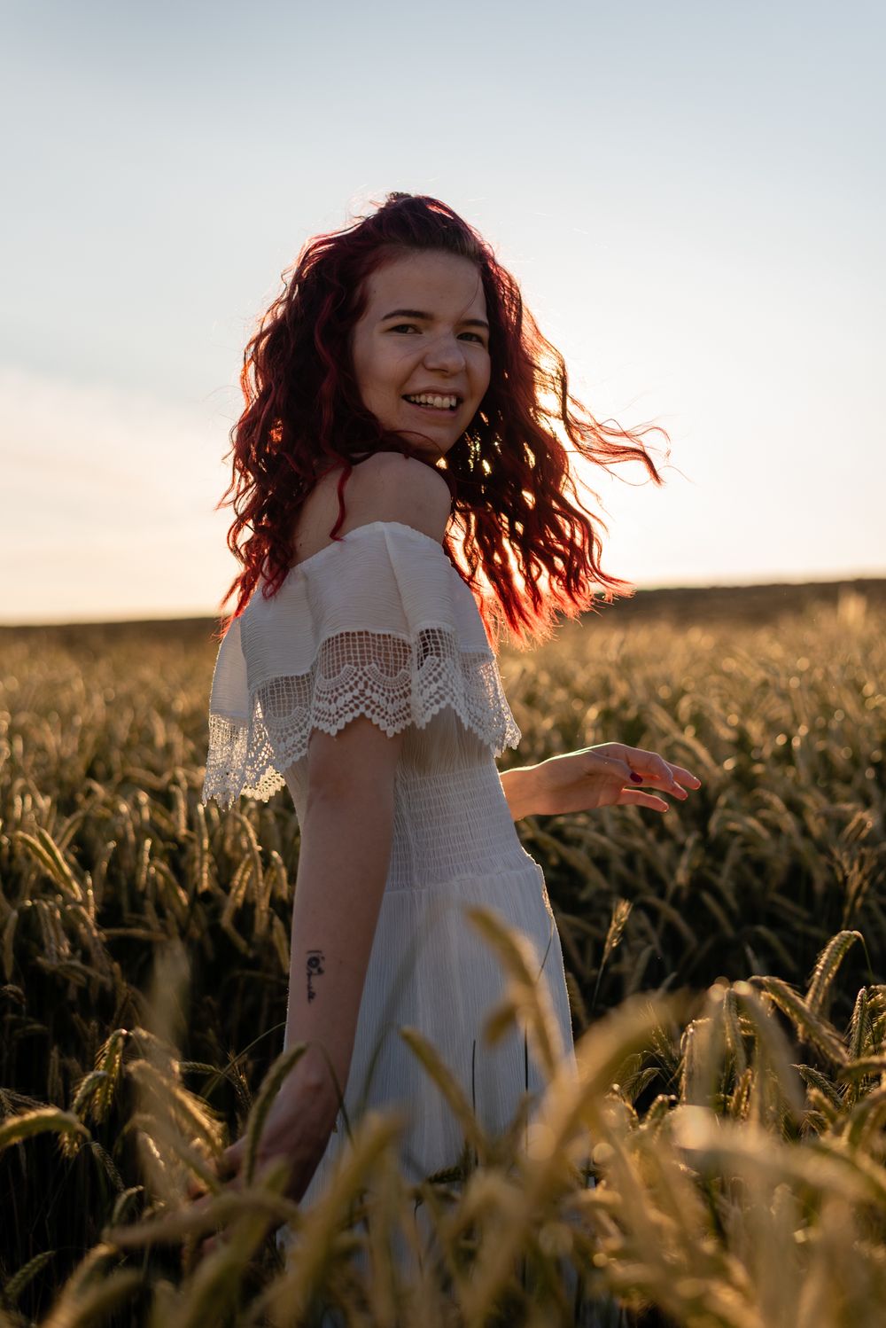 Happy young woman with red curly hair wearing white off-shoulder dress in golden wheat field at sunset.