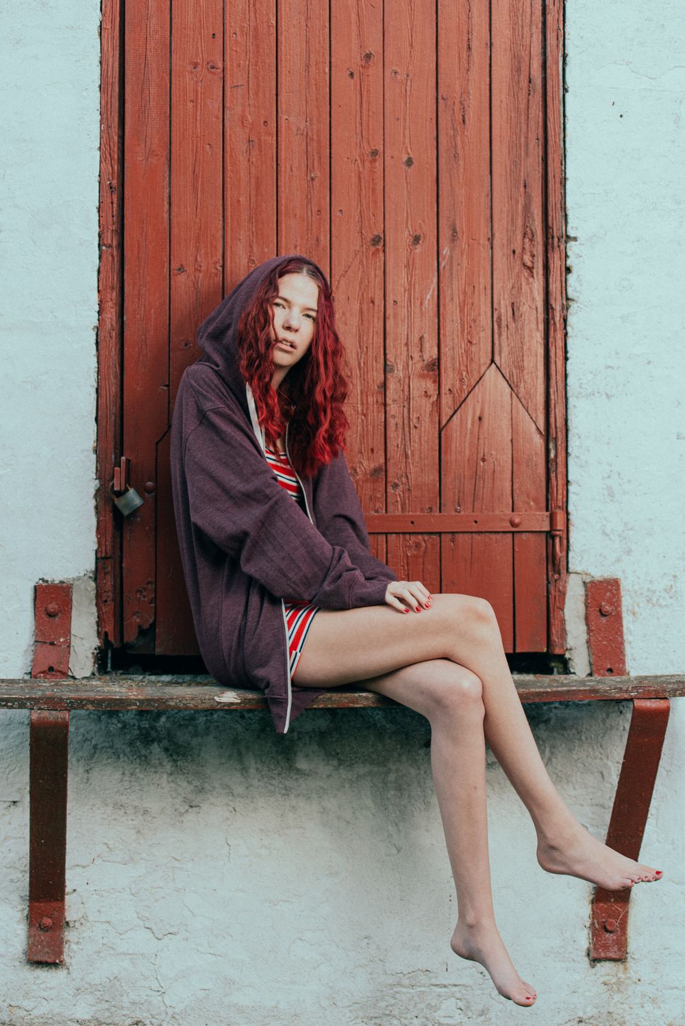 Young woman with red curly hair sitting barefoot on ledge against rustic red wooden door.