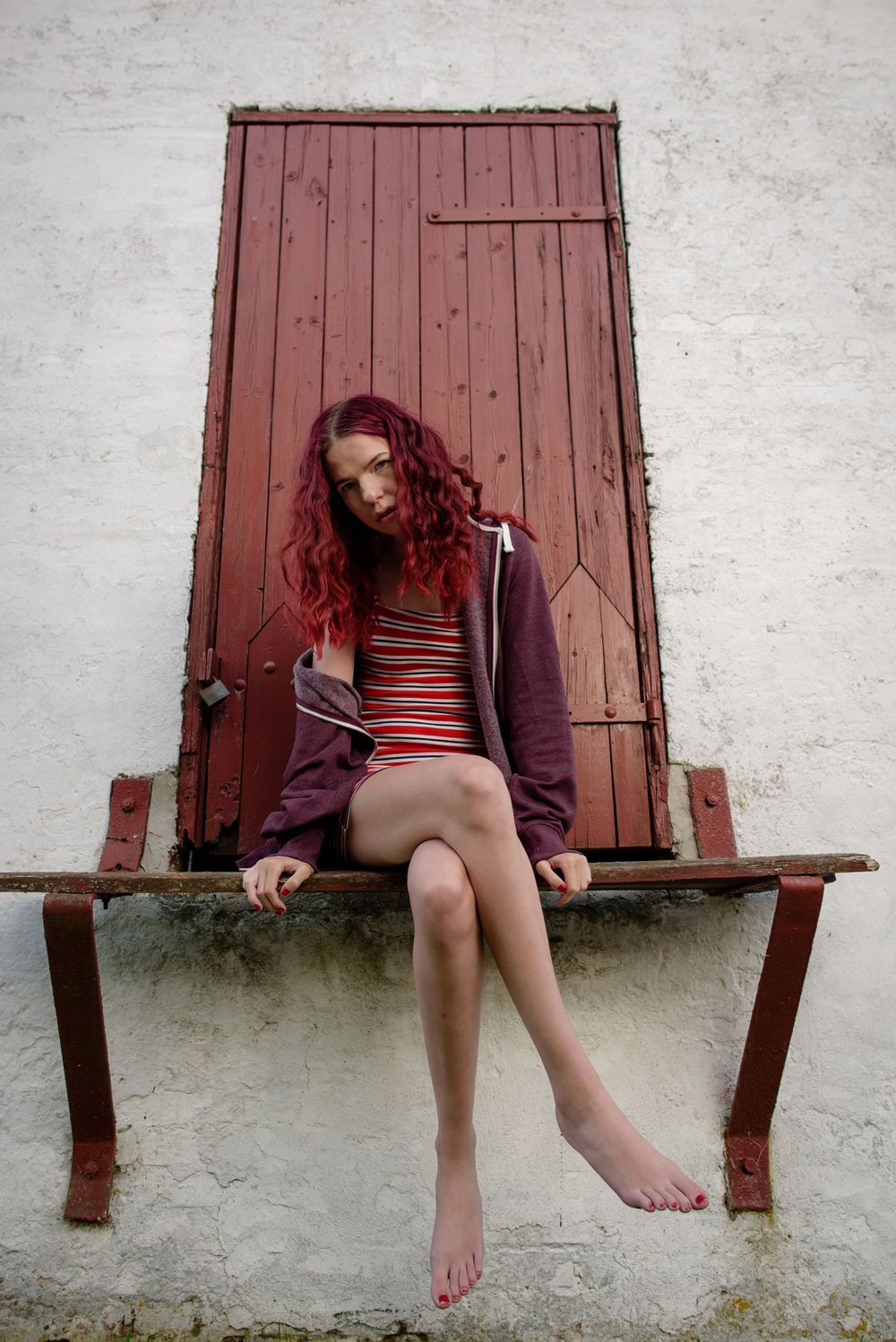 Red-haired woman in striped dress and hoodie sitting barefoot on metal ledge by red door.