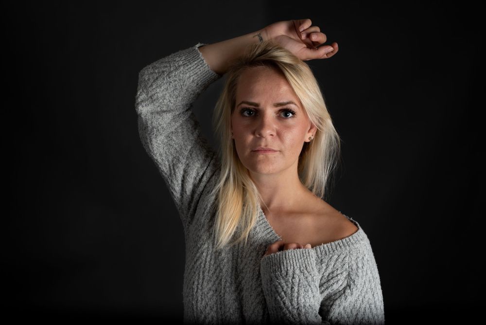 Blonde woman in gray off-shoulder sweater posing with arm raised against dark studio background.