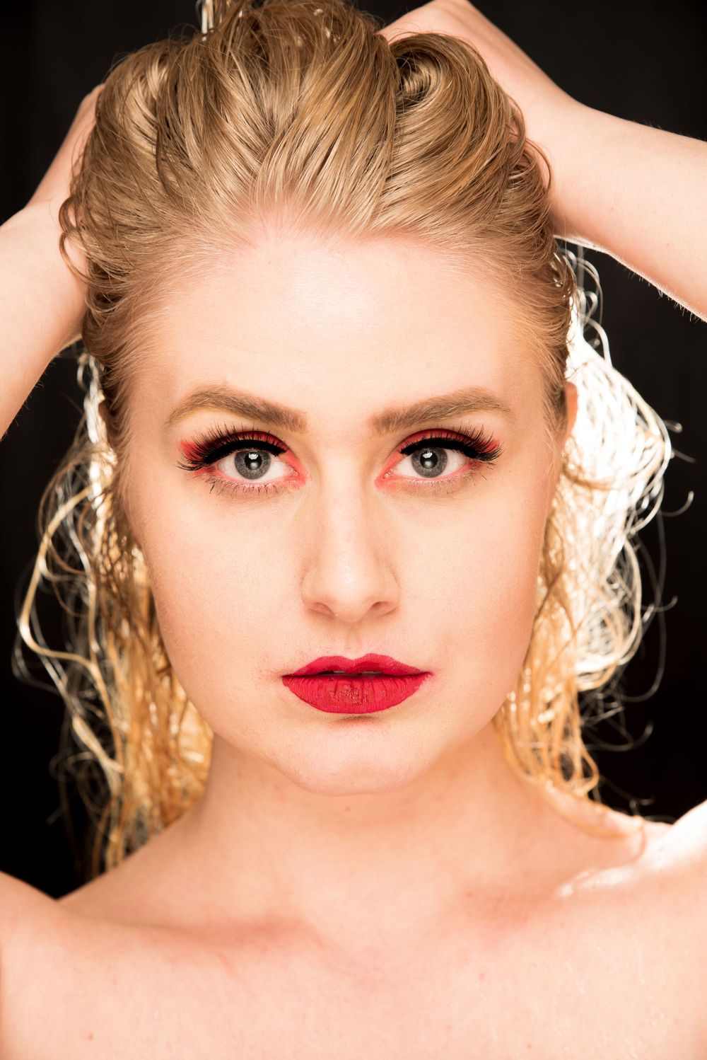 Blonde woman with bold red lips and dramatic eye makeup holding hair up against black studio background.
