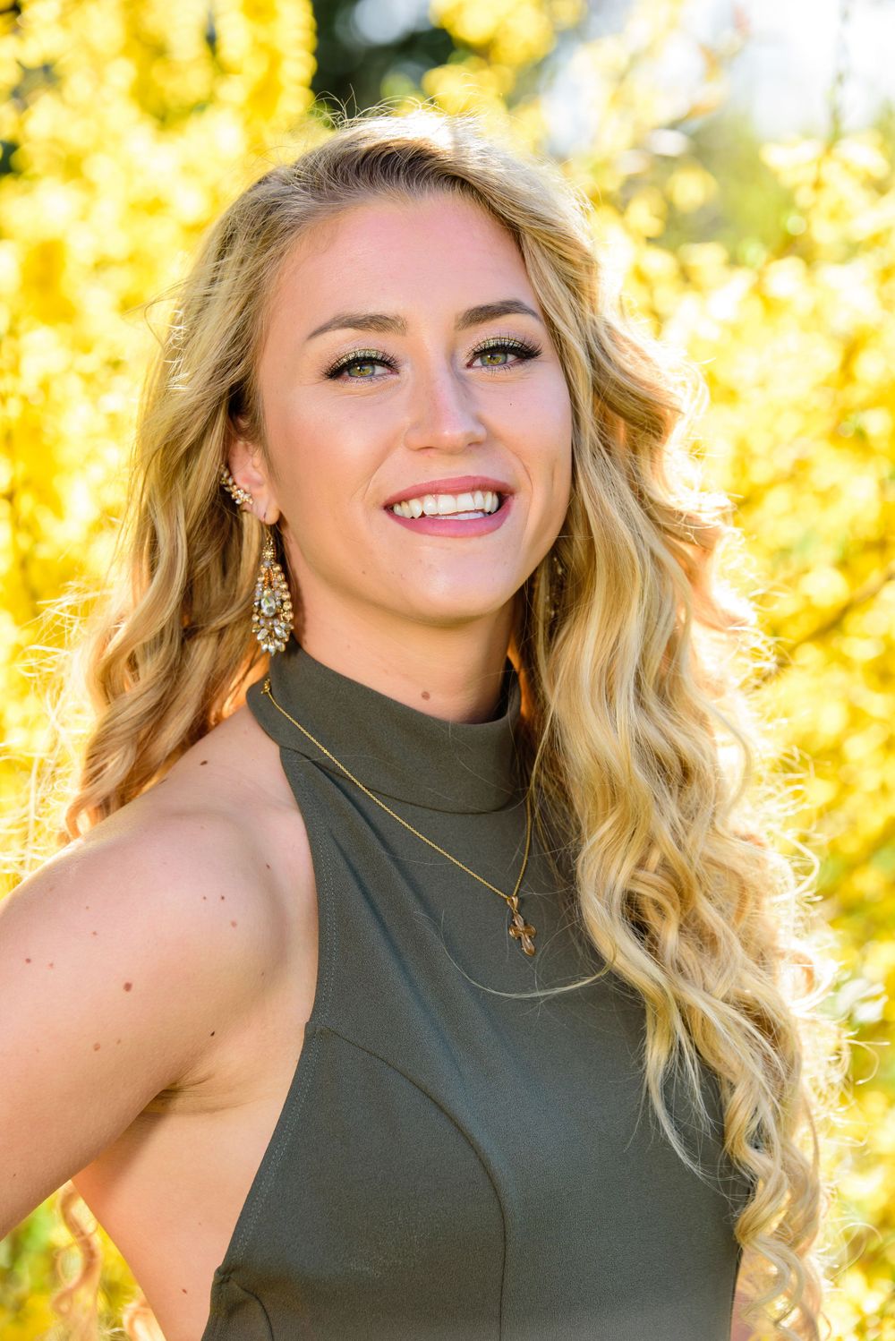 Smiling blonde woman with curly hair wearing olive green dress and cross necklace against yellow autumn foliage.