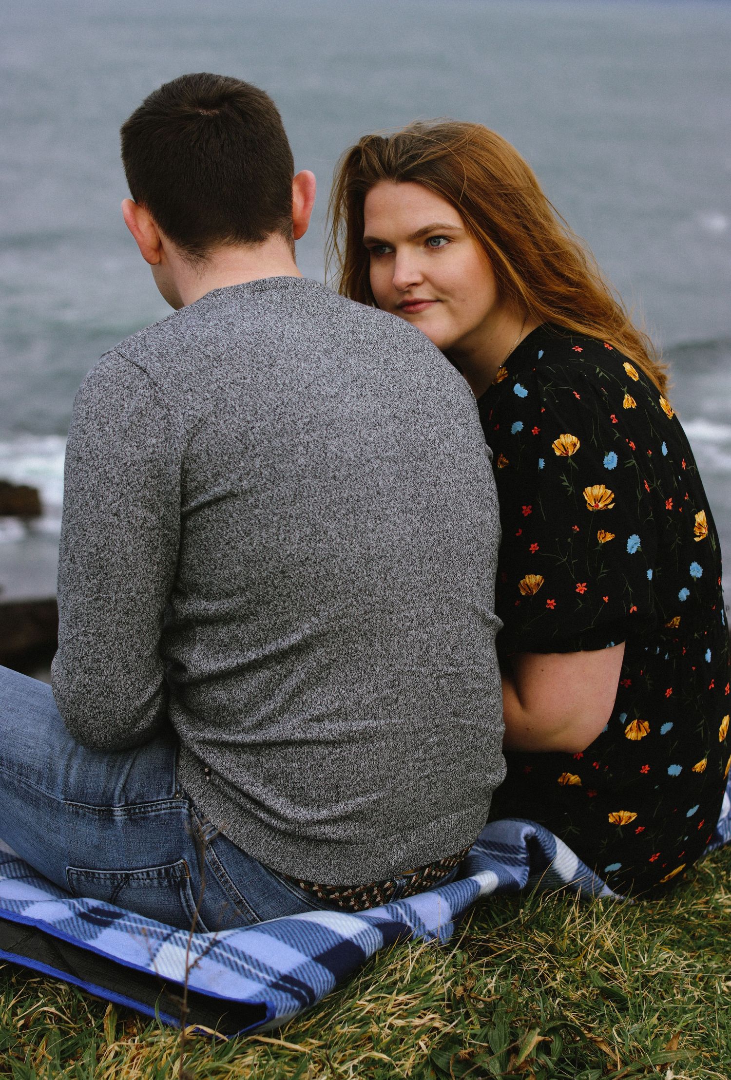 Couple sitting on a blanket by the ocean as woman in floral dress looks at camera.