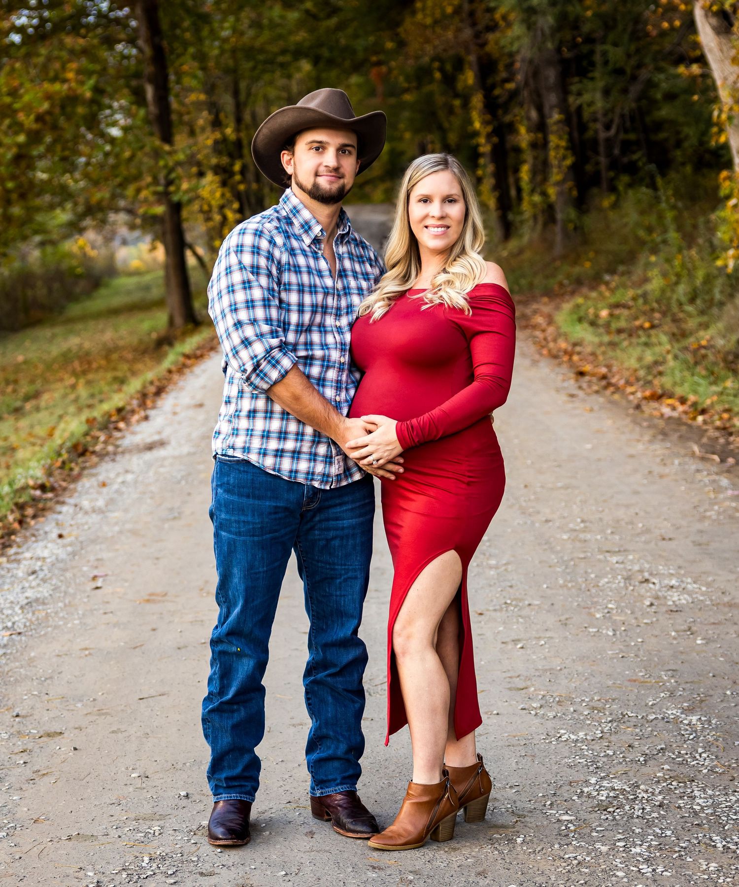 Maternity photoshoot of couple on autumn path with woman in red dress and man in plaid shirt.