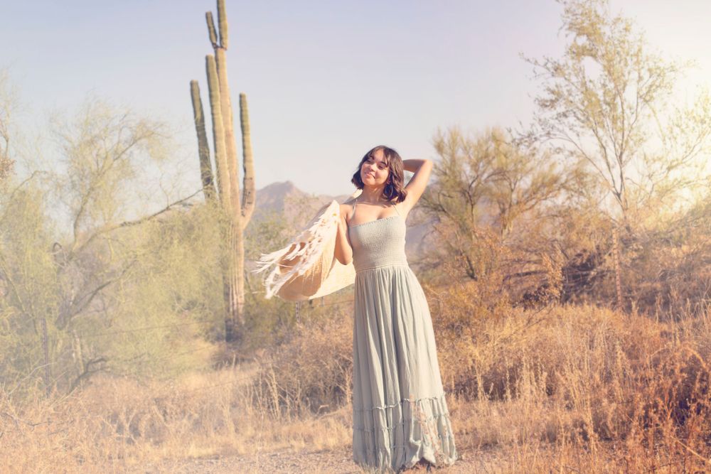 Woman in flowing dress poses gracefully by saguaro cactus in desert landscape.