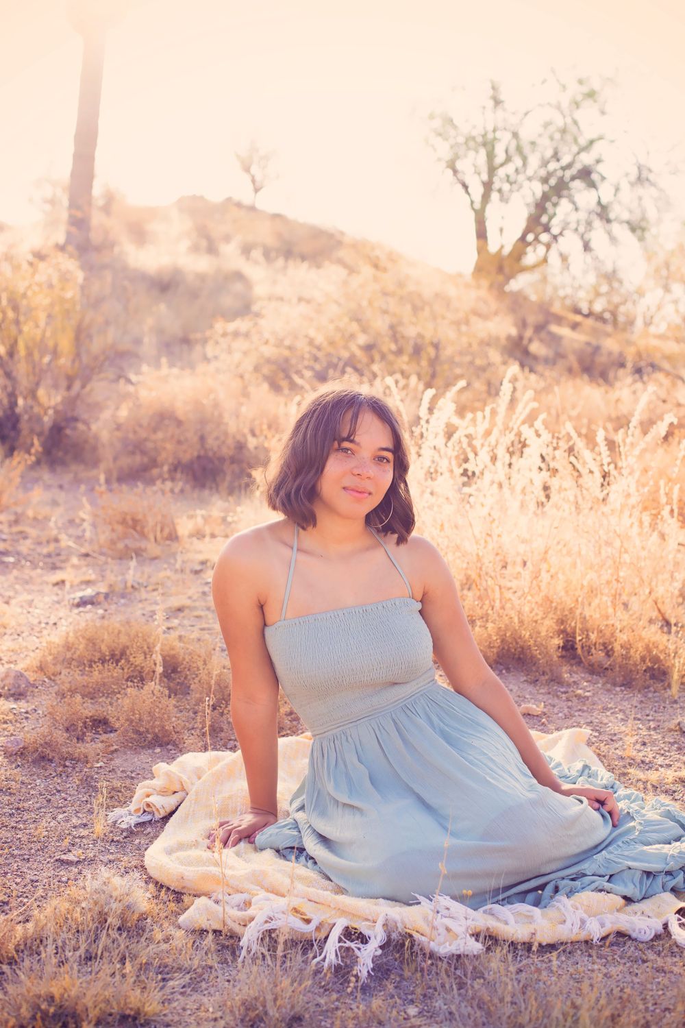 Woman in light blue dress sits on blanket in desert setting during sunset.