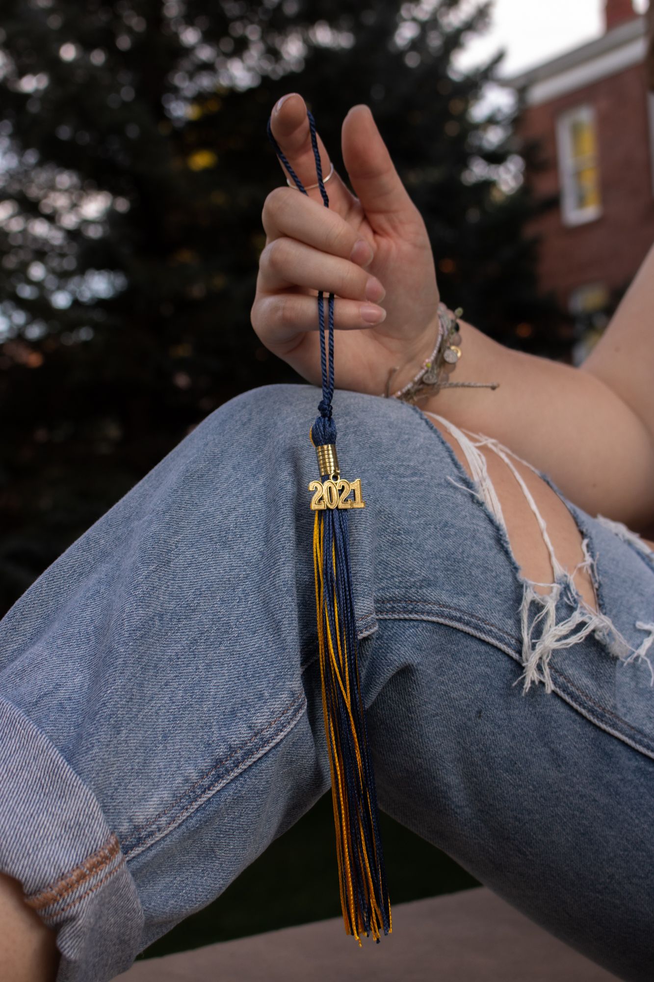 Hand holding black and gold graduation tassel with 2021 charm while sitting in ripped jeans.