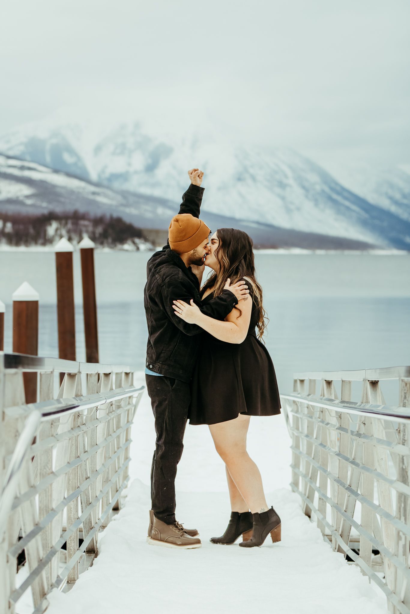 A romantic winter embrace on a snowy dock overlooking snow-capped mountains and a serene lake.