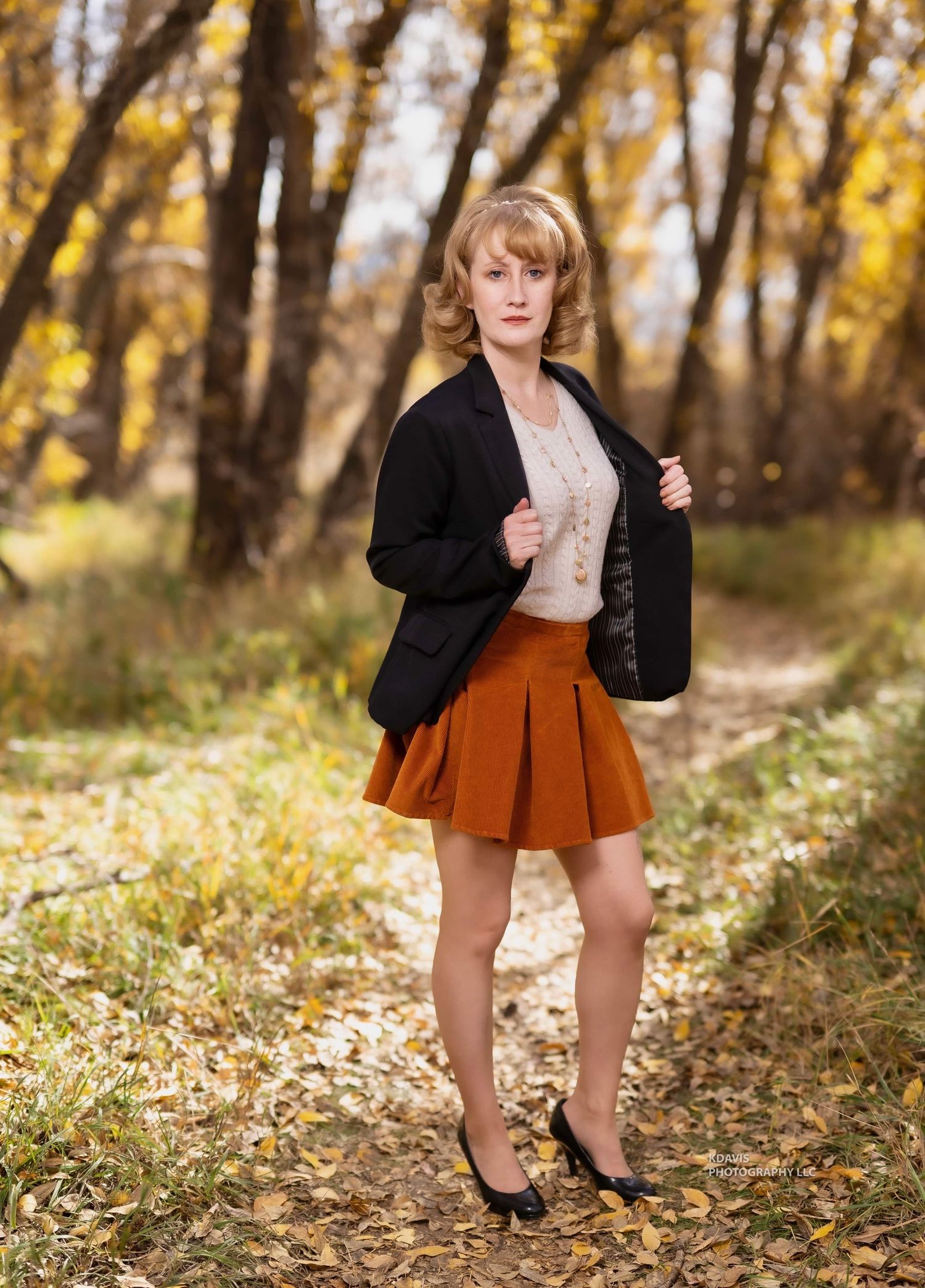 Image of Mon Ami Mindy in vintage-inspired autumn outfit featuring an orange pleated skirt and black blazer in a wooded setting.