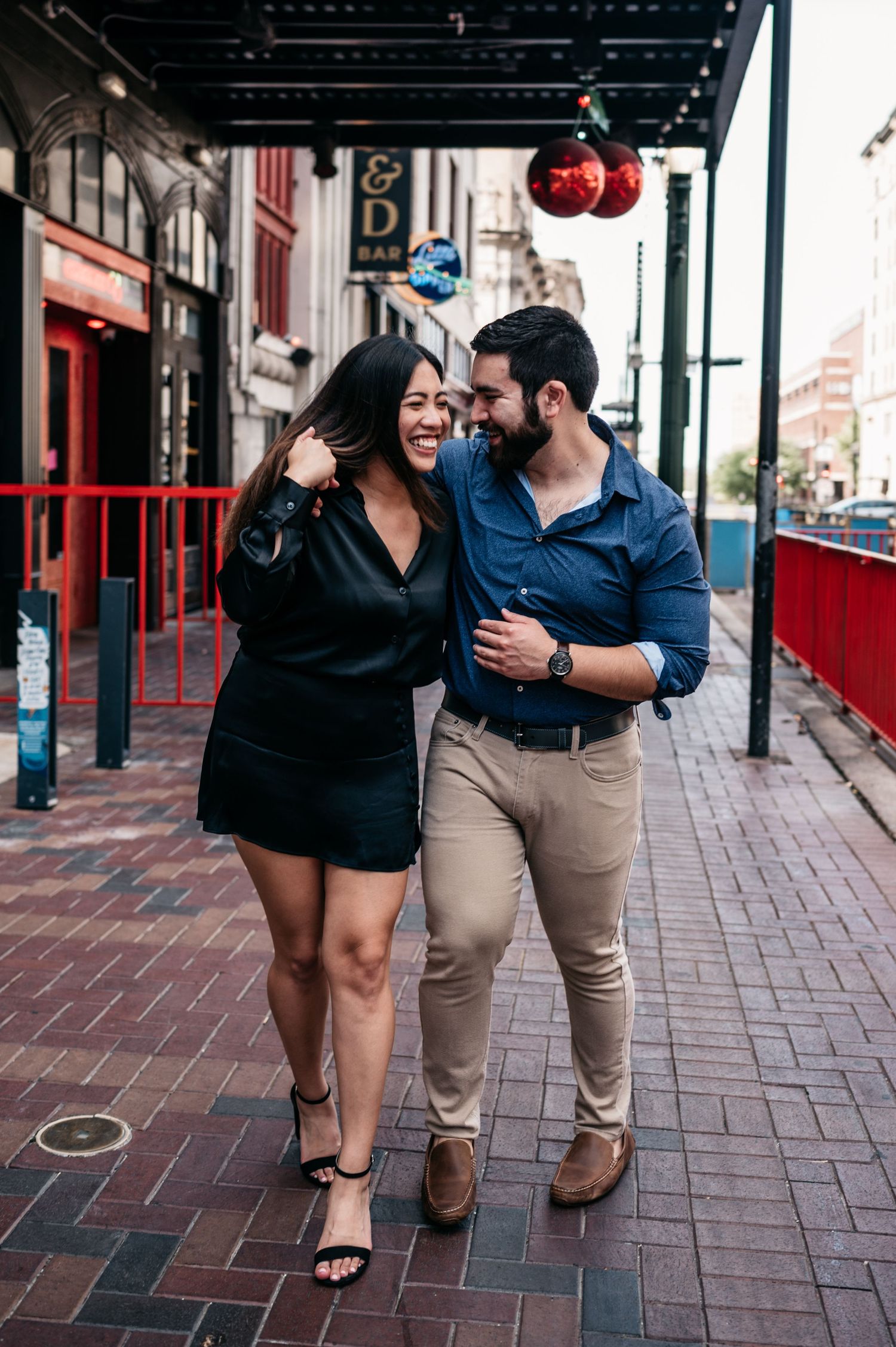 A couple shares a laugh while walking under decorative ornaments on a brick sidewalk.