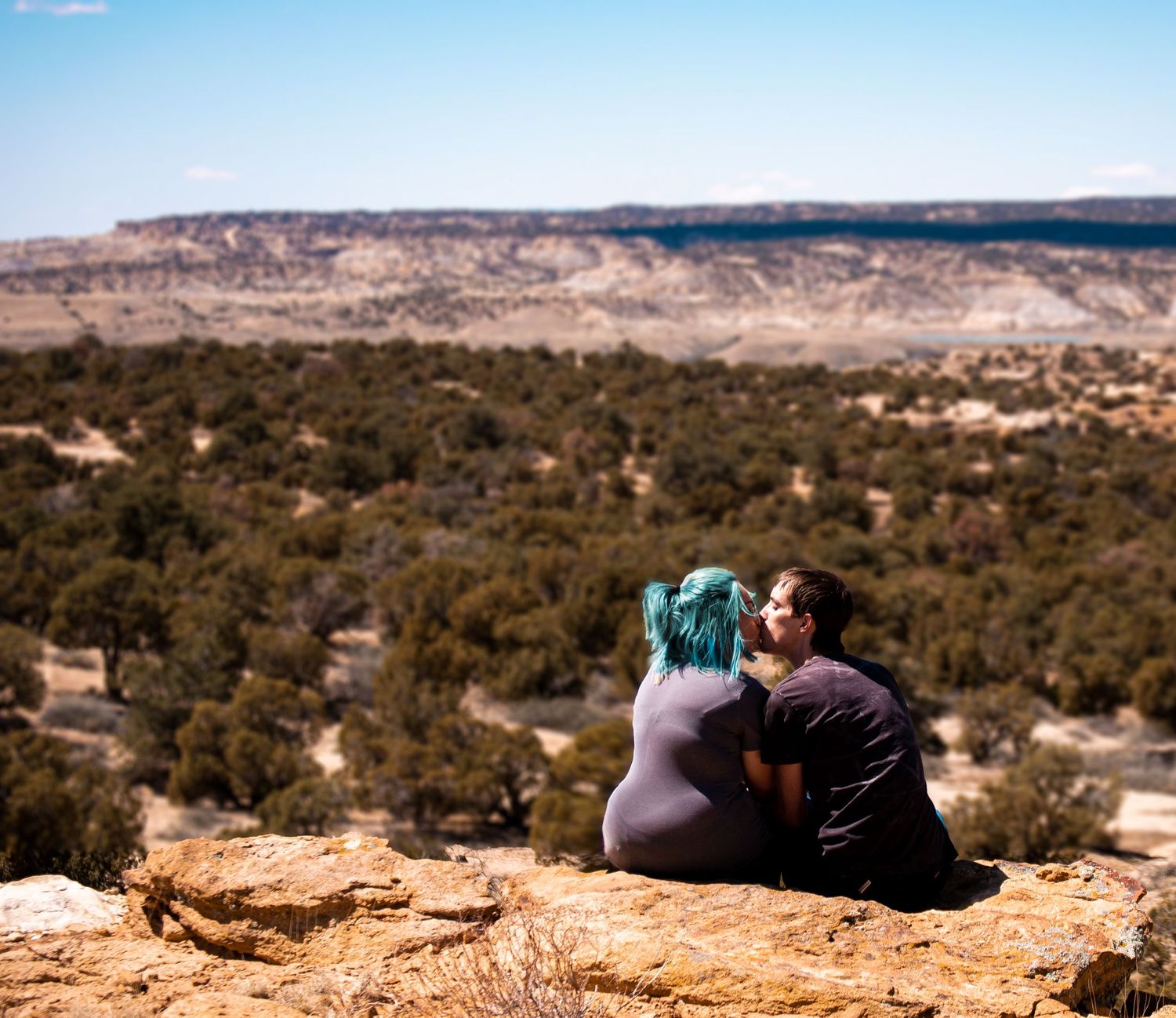 A couple kisses on a mountain cliffside, with a distant mesa in the background.