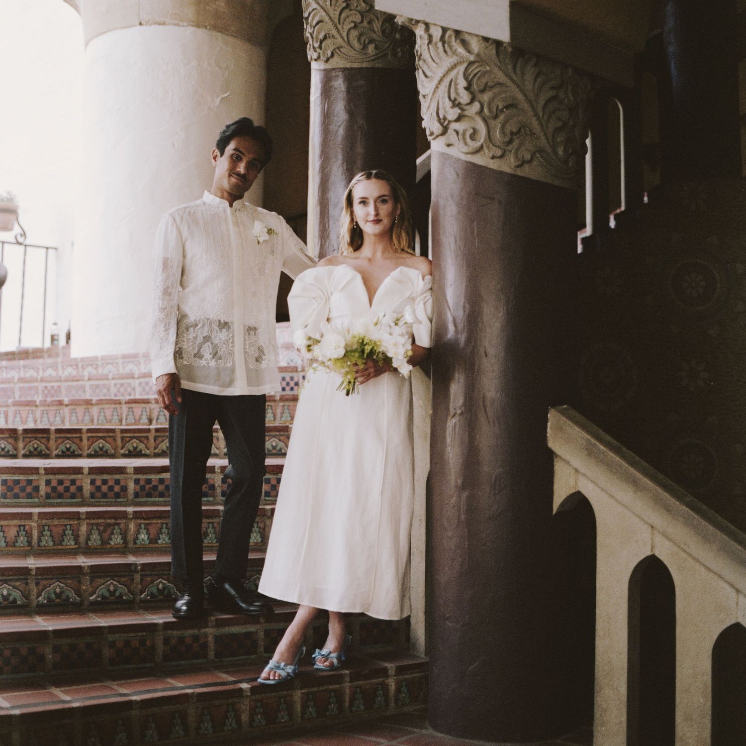 Newlyweds pose on decorative tiled stairs against ornate architectural columns.