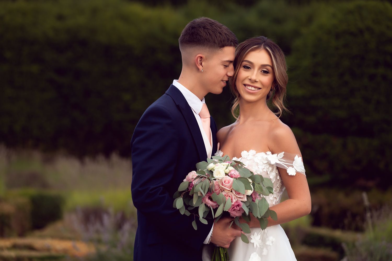 A couple shares an intimate moment in their wedding attire while holding a pink and white bouquet.