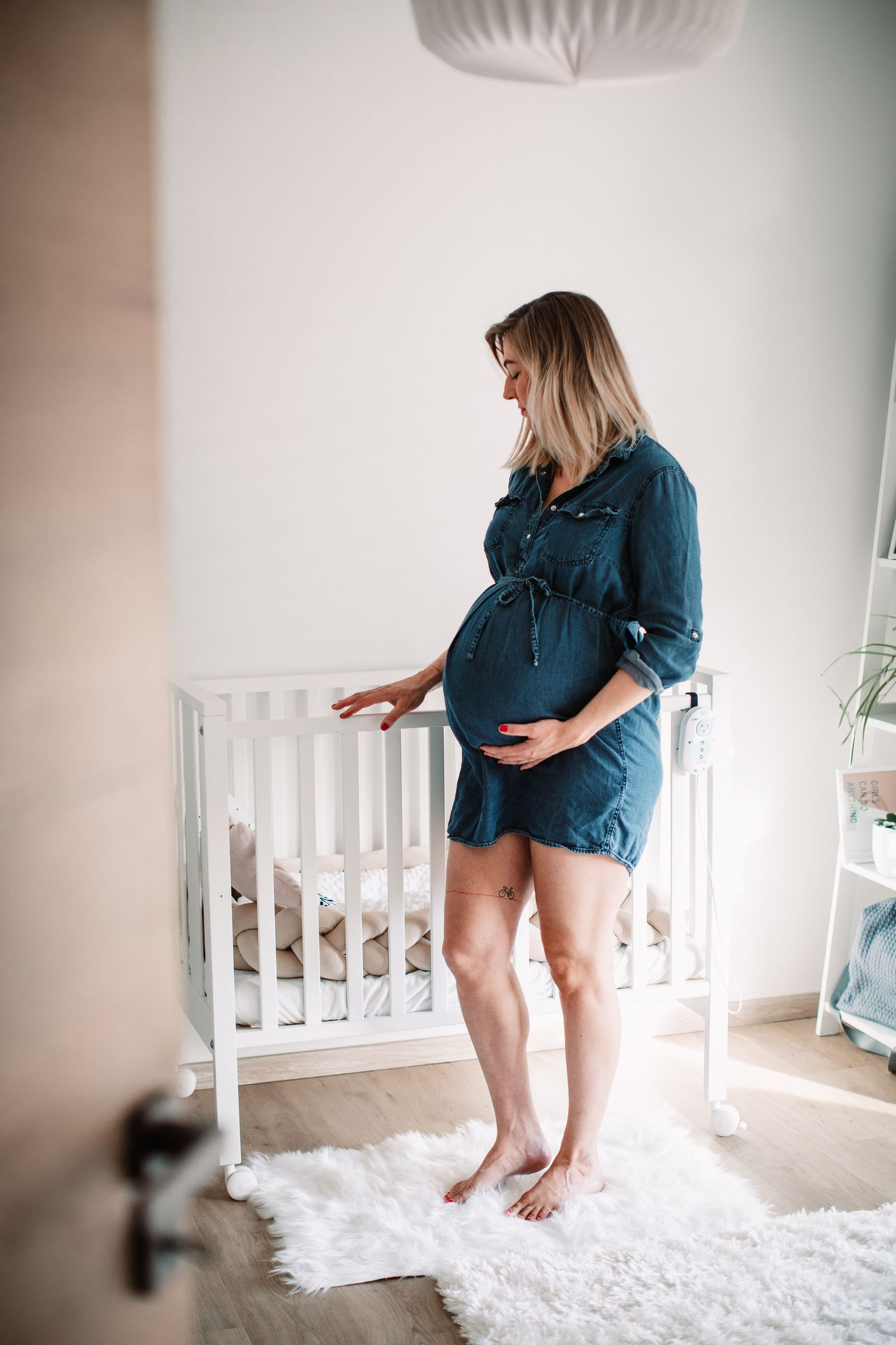 A pregnant woman in denim dress stands beside a white crib in a nursery.