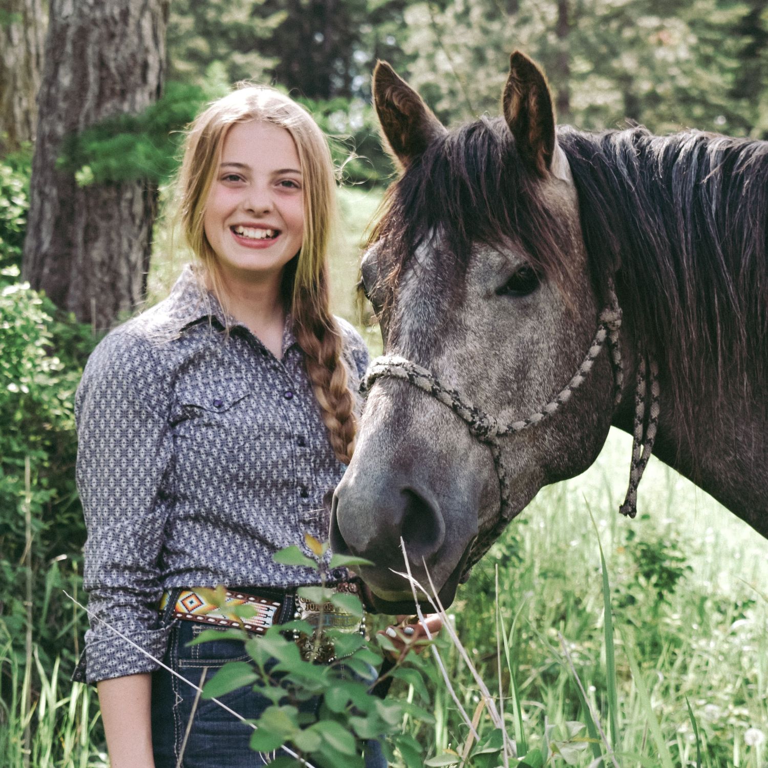 A dappled gray horse poses with someone in a button-down shirt against a forested background.