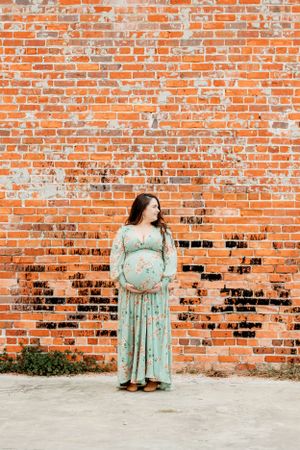A maternity photoshoot against a rustic red brick wall featuring a mint floral dress.