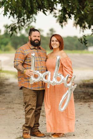 Pregnancy announcement photoshoot with couple holding 'baby' text sign outdoors.