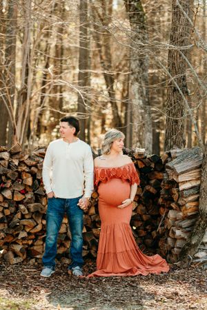 Rustic maternity photo of couple by woodpile in forest wearing white shirt and flowing rust-colored dress.