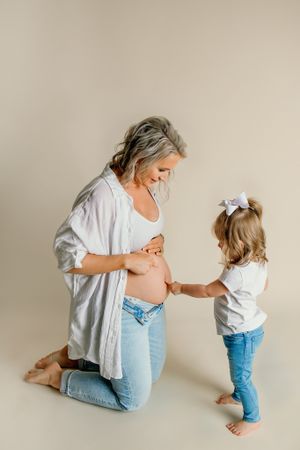 Sweet maternity moment with young child touching pregnant belly against neutral background.