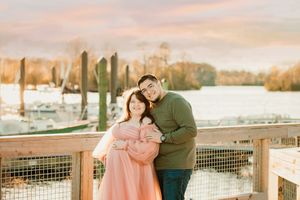 Expecting couple shares tender moment at marina dock during sunset in pink dress and green sweater.