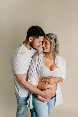 Intimate maternity portrait of couple in white and denim against neutral background.