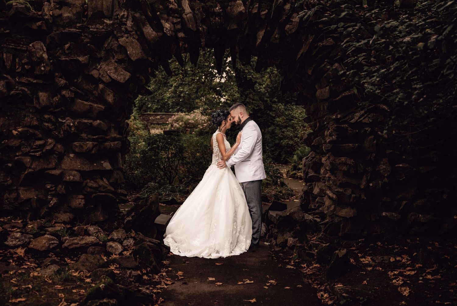 Romantic wedding photo of couple embracing under stone archway covered in autumn leaves.