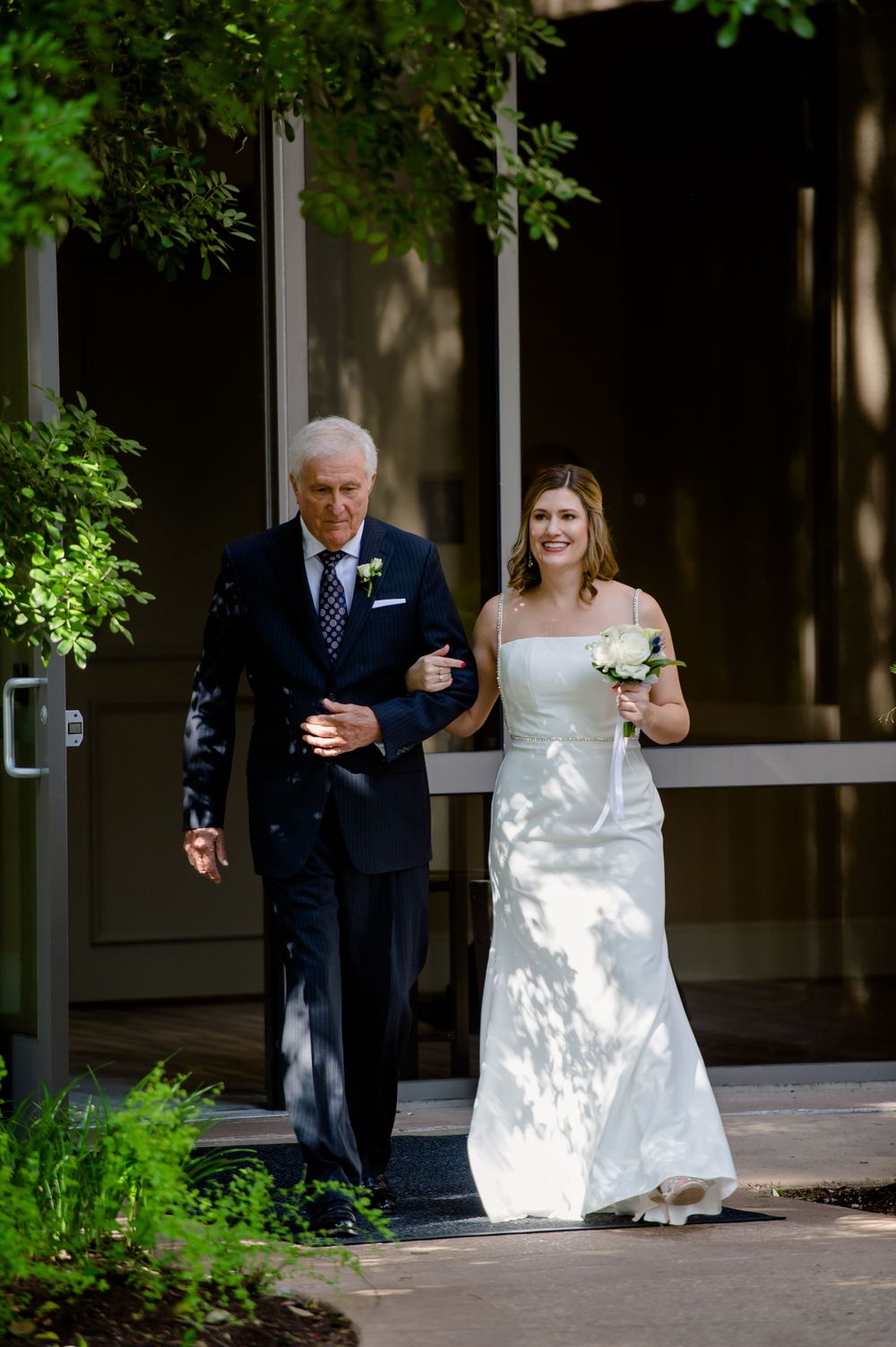 A pair walking arm in arm down a sunlit walkway lined with greenery.