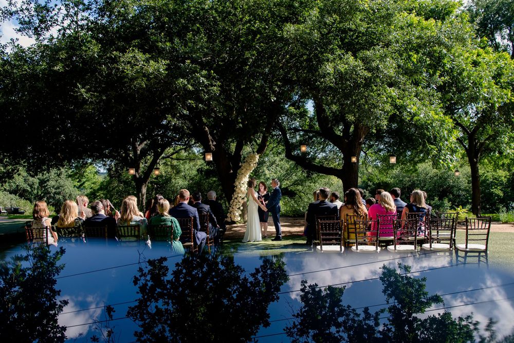 An outdoor wedding ceremony setup under large shade trees with white chairs.