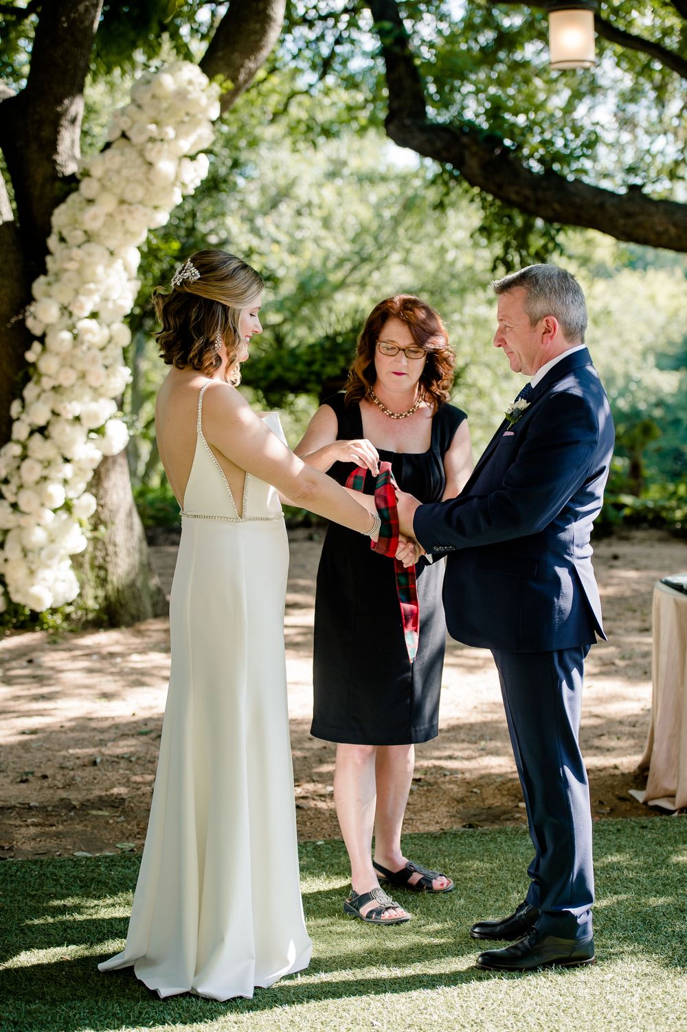 An intimate outdoor wedding ceremony under trees decorated with white floral arrangements.